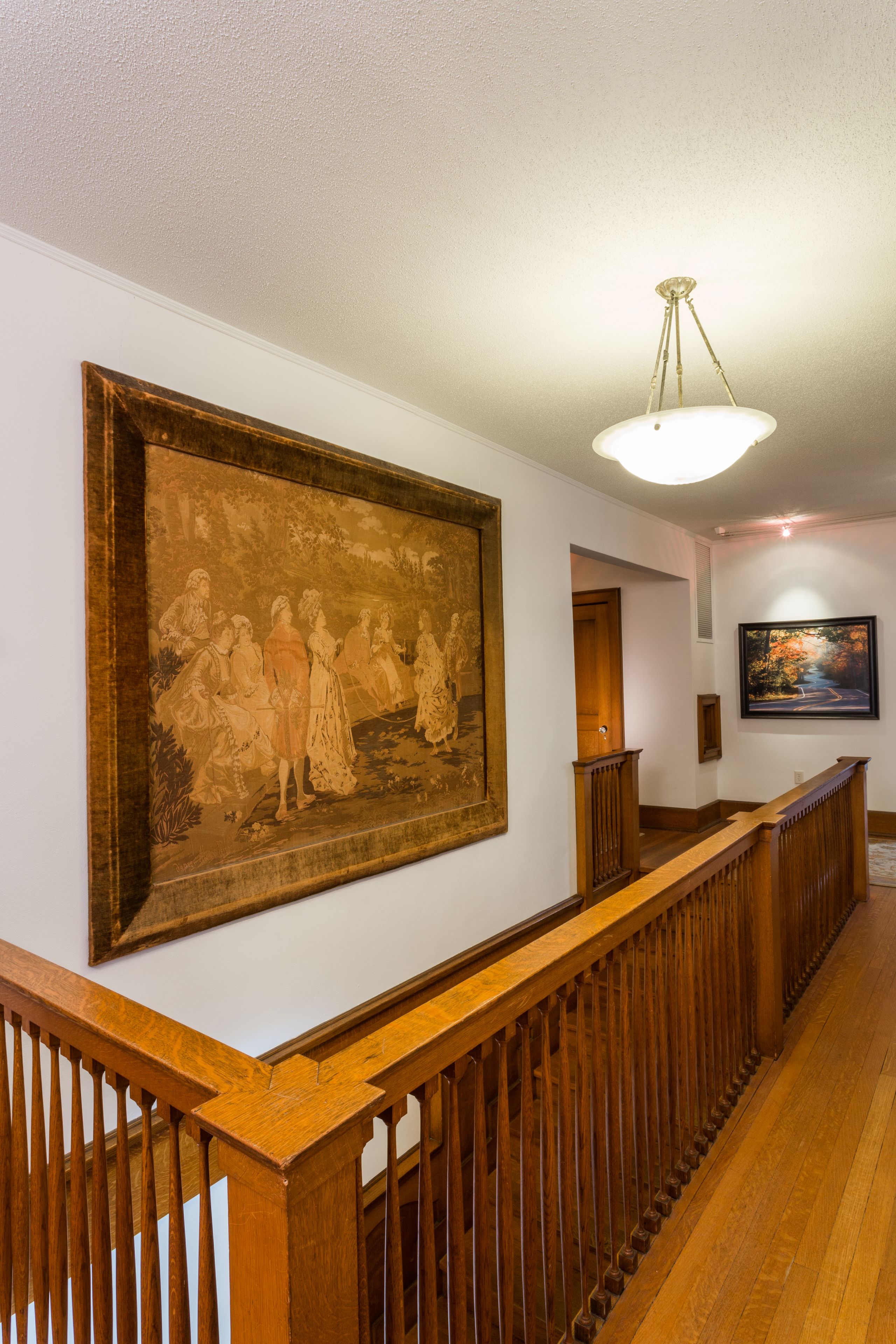 A hallway with a wooden railing, a large framed painting on the wall, and a ceiling light fixture.