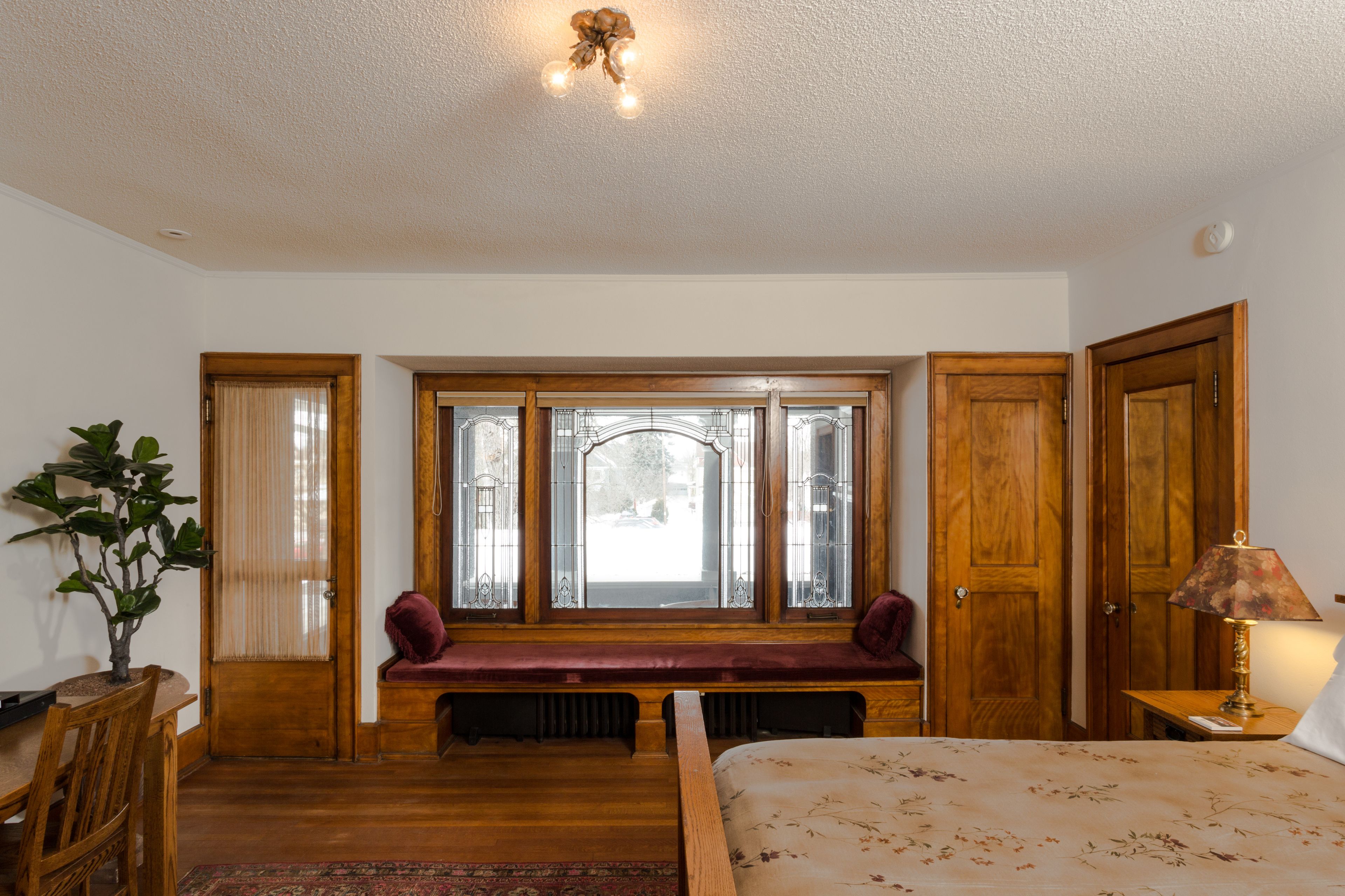 A bedroom with a wooden bench by the window, a bed with a floral bedspread, and a lamp on a bedside table.