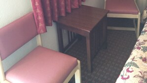 Um quarto pequeno com duas cadeiras de madeira, uma mesa, cortinas vermelhas e toalha de mesa estampada.