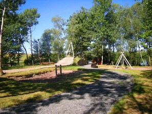 Explore a serene natural setting with a wooden bridge, swings, and a slide amidst lush trees and open grassy spaces.