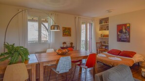 A cozy room with a wooden dining table, a bed with red pillows, a bookshelf, and a large window with curtains.