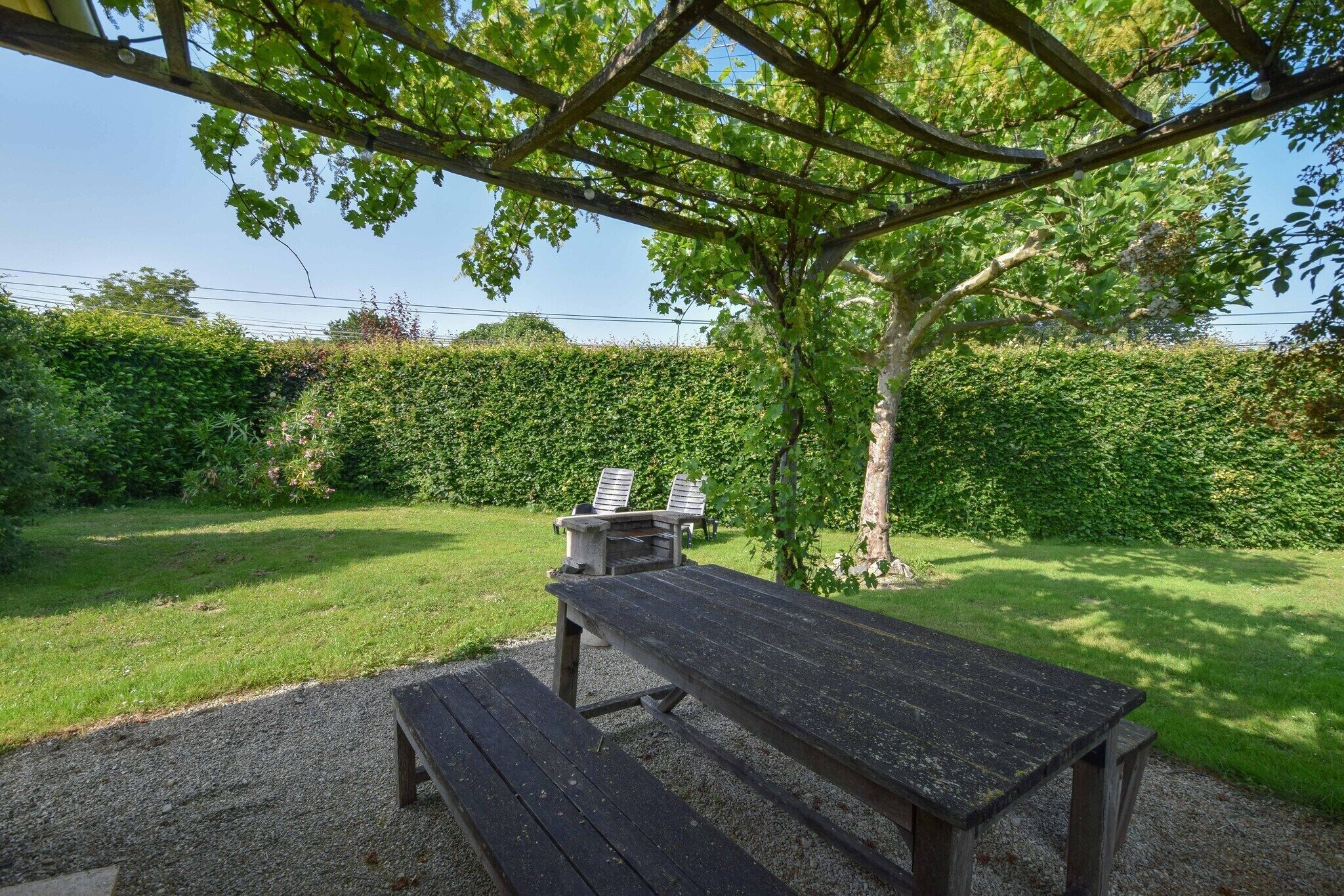 An outdoor seating area with a wooden table and benches, surrounded by a hedge and trees.