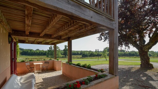 A covered outdoor seating area with a table and chairs, a wooden pergola, and a view of a green landscape.