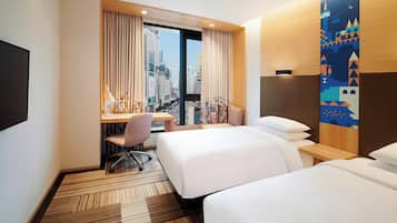 Habitación de hotel con dos camas, un escritorio con silla, televisor y vistas a la ciudad a través de ventanas amplias.