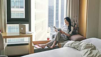 Une chambre dâhĂŽtel avec un lit, un bureau avec un ordinateur portable, une fenĂȘtre donnant sur des immeubles imposants, et une personne lisant un livre.