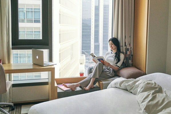 A hotel room with a bed, a desk with a laptop, a window with a view of tall buildings, and a person reading a book.