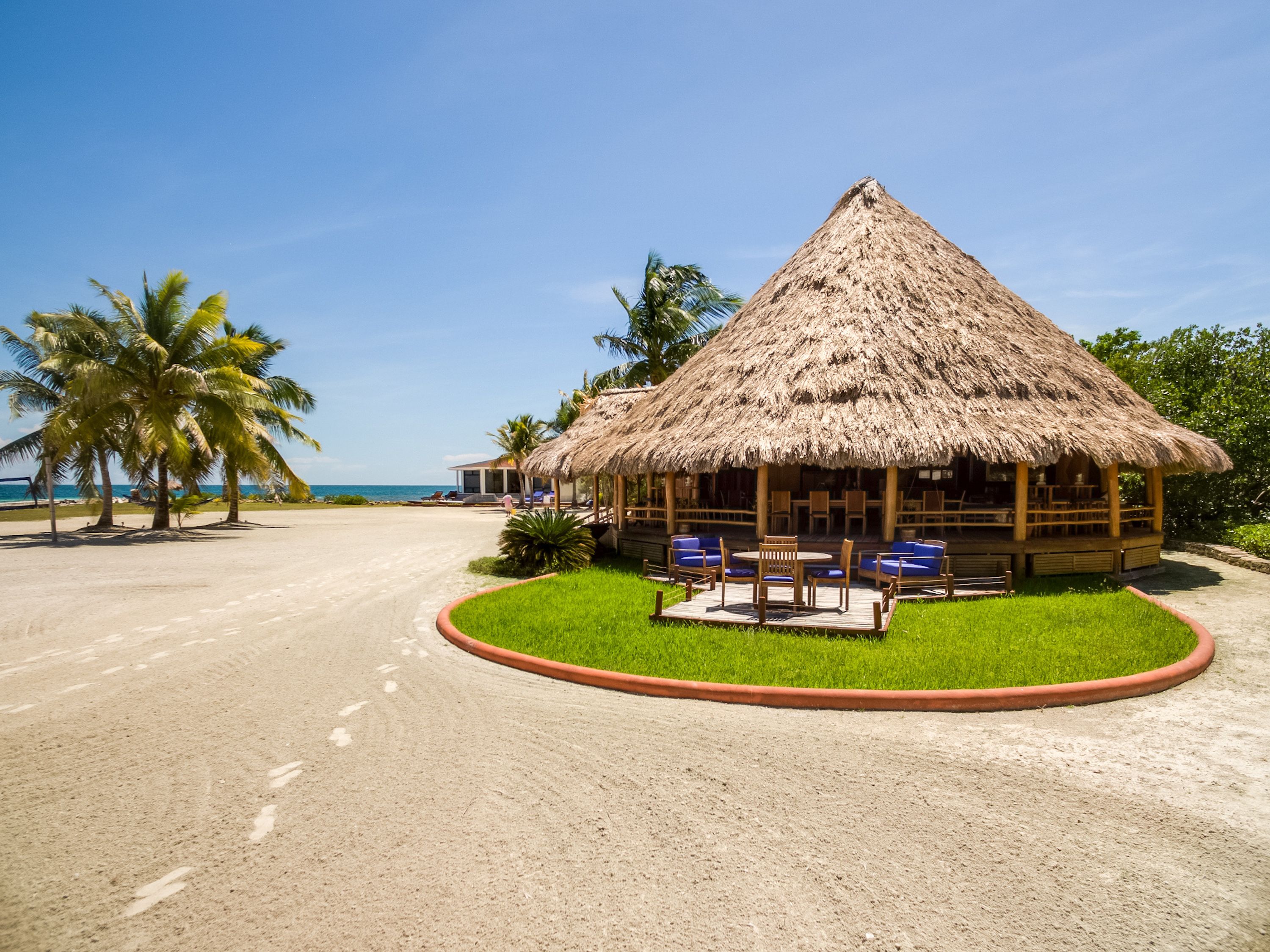 Una cabaña con techo de paja y mobiliario exterior, rodeada de palmeras y una playa de arena.