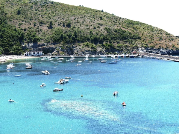 Une zone côtière avec des bateaux sur une eau bleu clair, une colline et un bâtiment à la façade blanche.