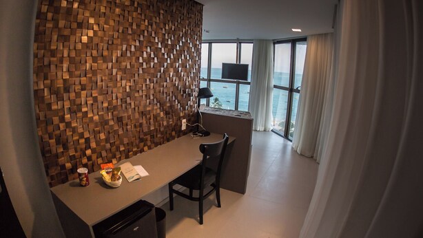 A modern hotel room with a wooden wall, a desk with a chair, a TV, and a view of the sea. - Acqua Suites Maceio (Maceió)