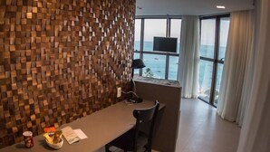 A modern hotel room with a wooden wall, a desk with a chair, a trash bin, a small table with snacks, and a TV. - Acqua Suites Maceio (Maceió)