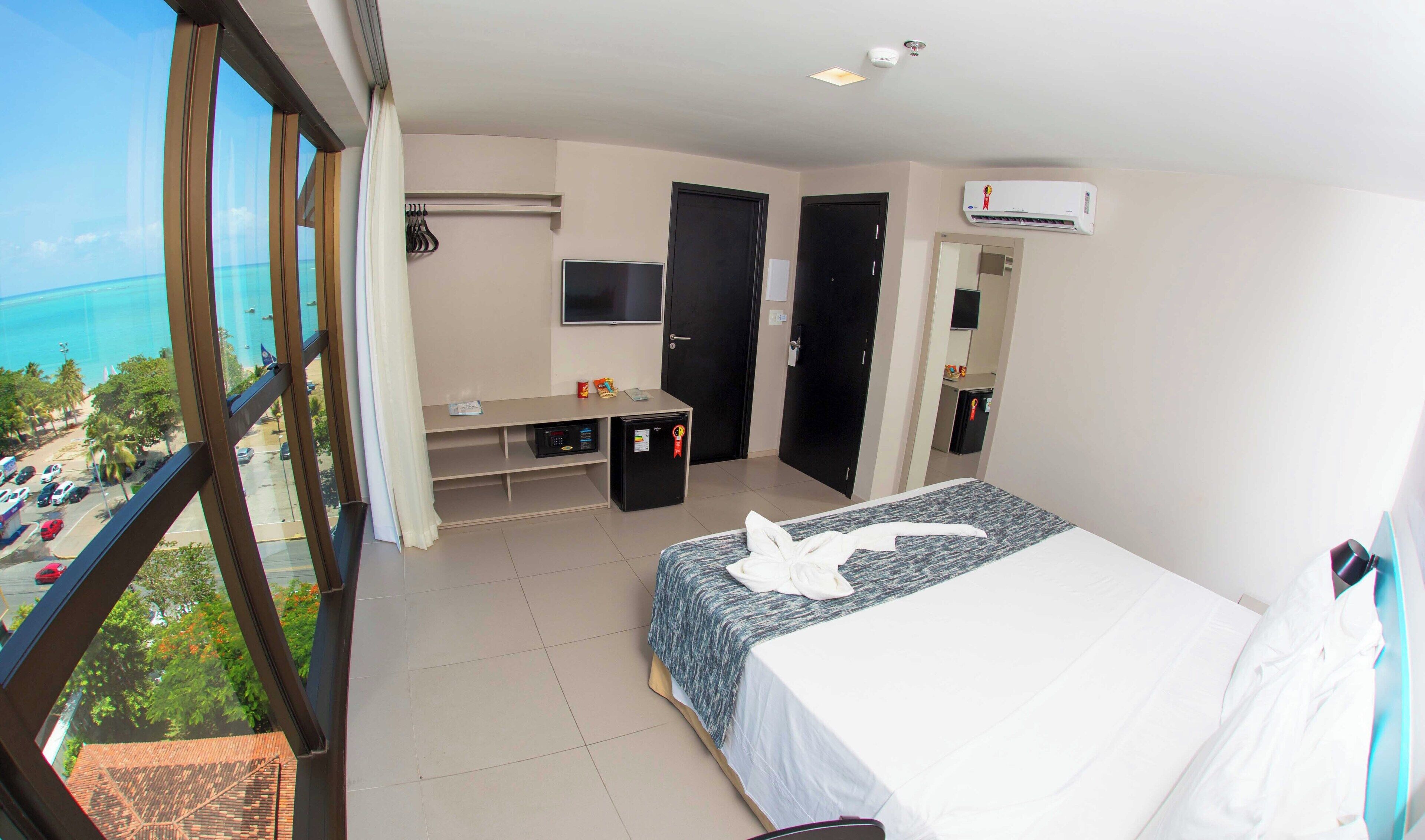 A hotel room with a bed, a TV, a mini-fridge, a desk, and a balcony with a view of the ocean.
