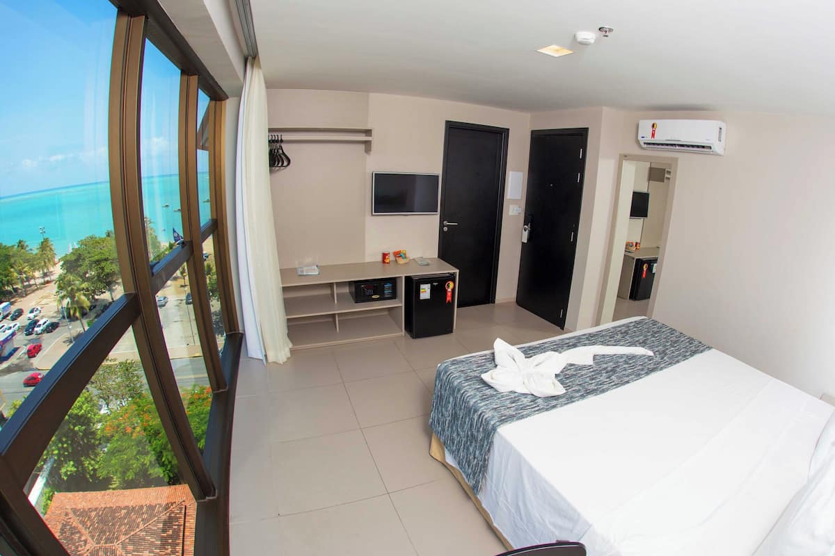 Une chambre d’hôtel équipée d’un lit, d’une télévision, d’un mini-réfrigérateur, d’un bureau et d’un balcon avec vue sur l’océan.