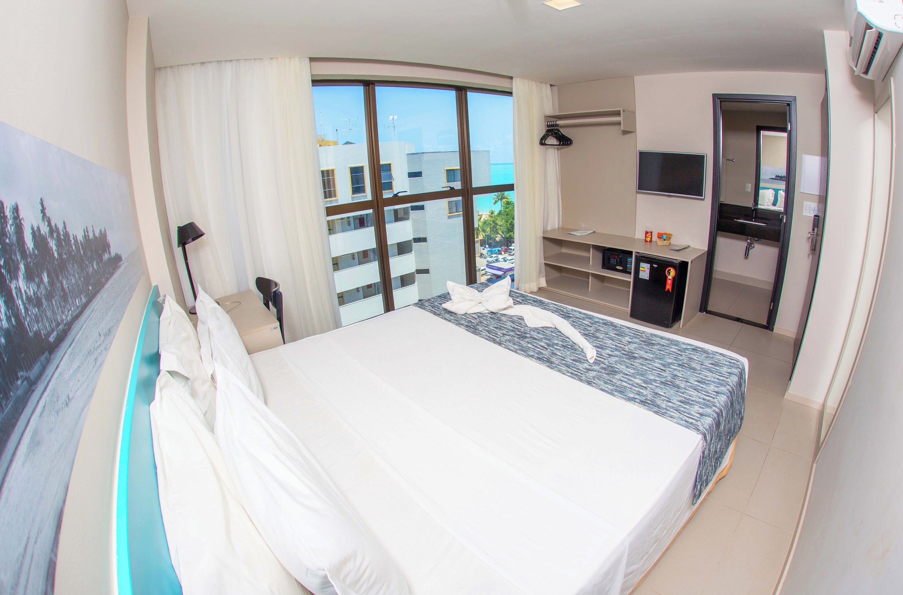 A hotel room with a large bed, a desk, a TV, and a view of the city through large windows.