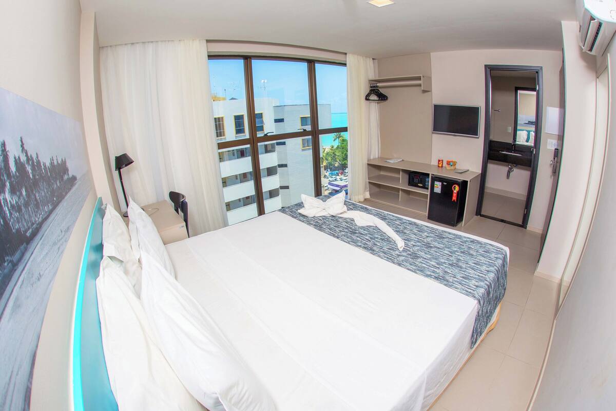 Une chambre d’hôtel avec un grand lit, un bureau, une télévision et une vue sur la ville grâce à de grandes fenêtres.