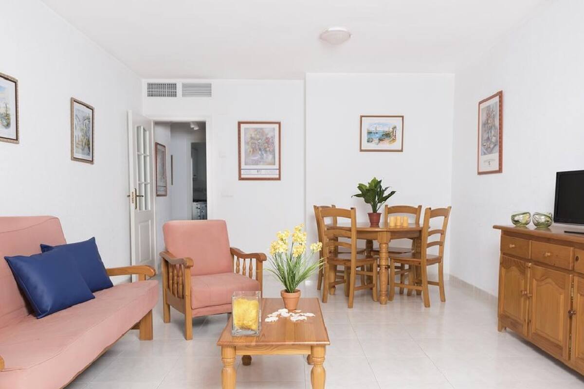 Un salon comprenant un canapé, un fauteuil, une table à manger en bois et des chaises. On y trouve également un téléviseur posé sur un meuble en bois, et des œuvres d’art accrochées aux murs.