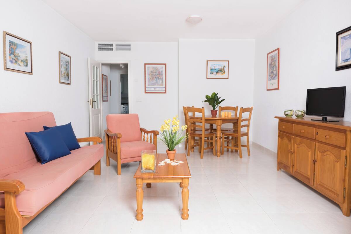 Un salon avec un canapé rose, une table basse en bois sur laquelle est posé un vase de fleurs, une table à manger avec des chaises, et un téléviseur placé sur un meuble en bois.