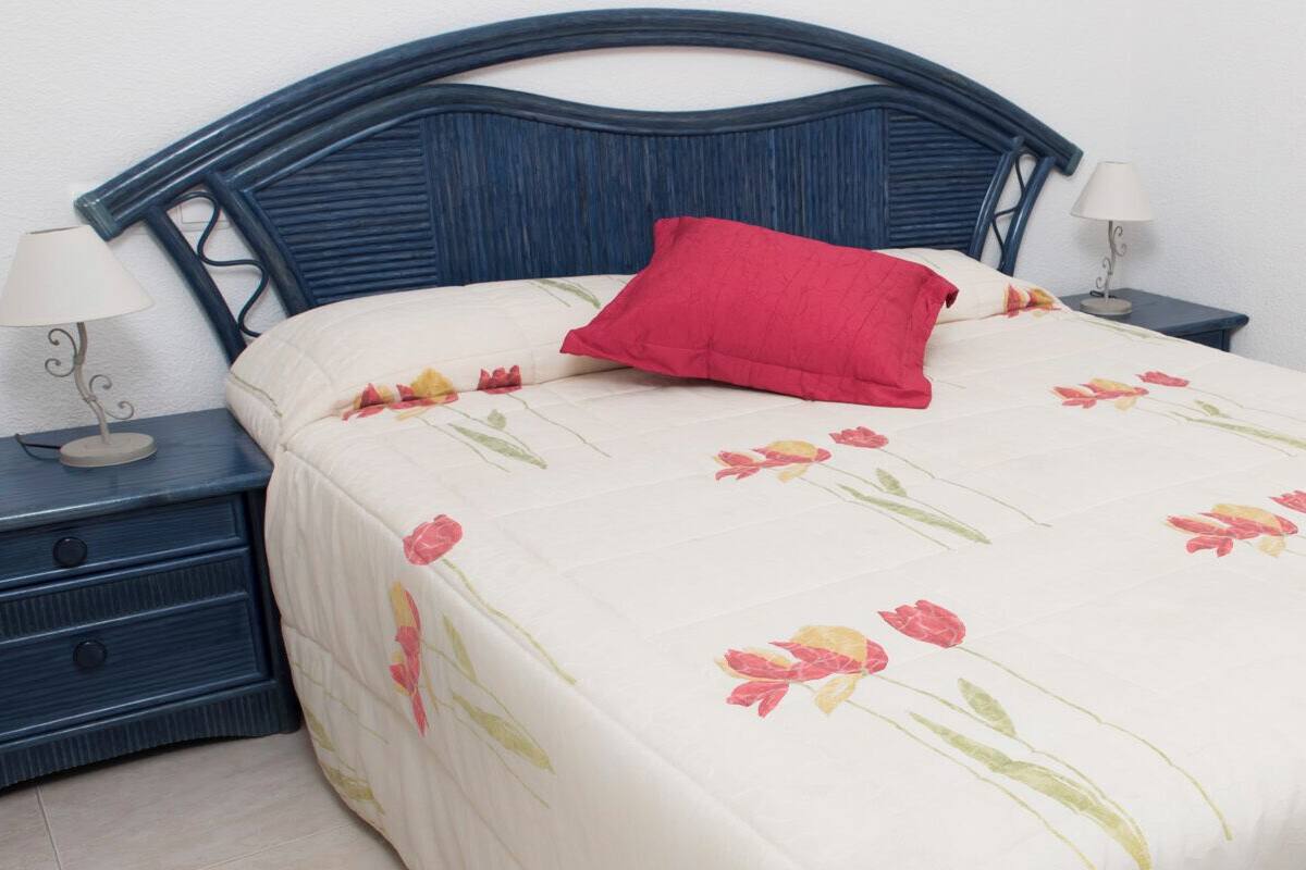 Un lit recouvert d’une couvre-lit à motifs floraux, un oreiller rouge et une tête de lit bleu foncé. Il y a deux tables de chevet avec des lampes et une petite table de nuit avec une lampe.