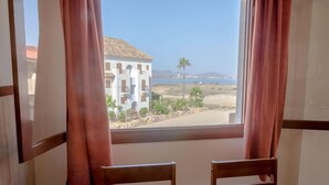 A room with a large window overlooking a beach, two wooden chairs, and a view of a building with a tiled roof. - Tesy II (LA MANGA)