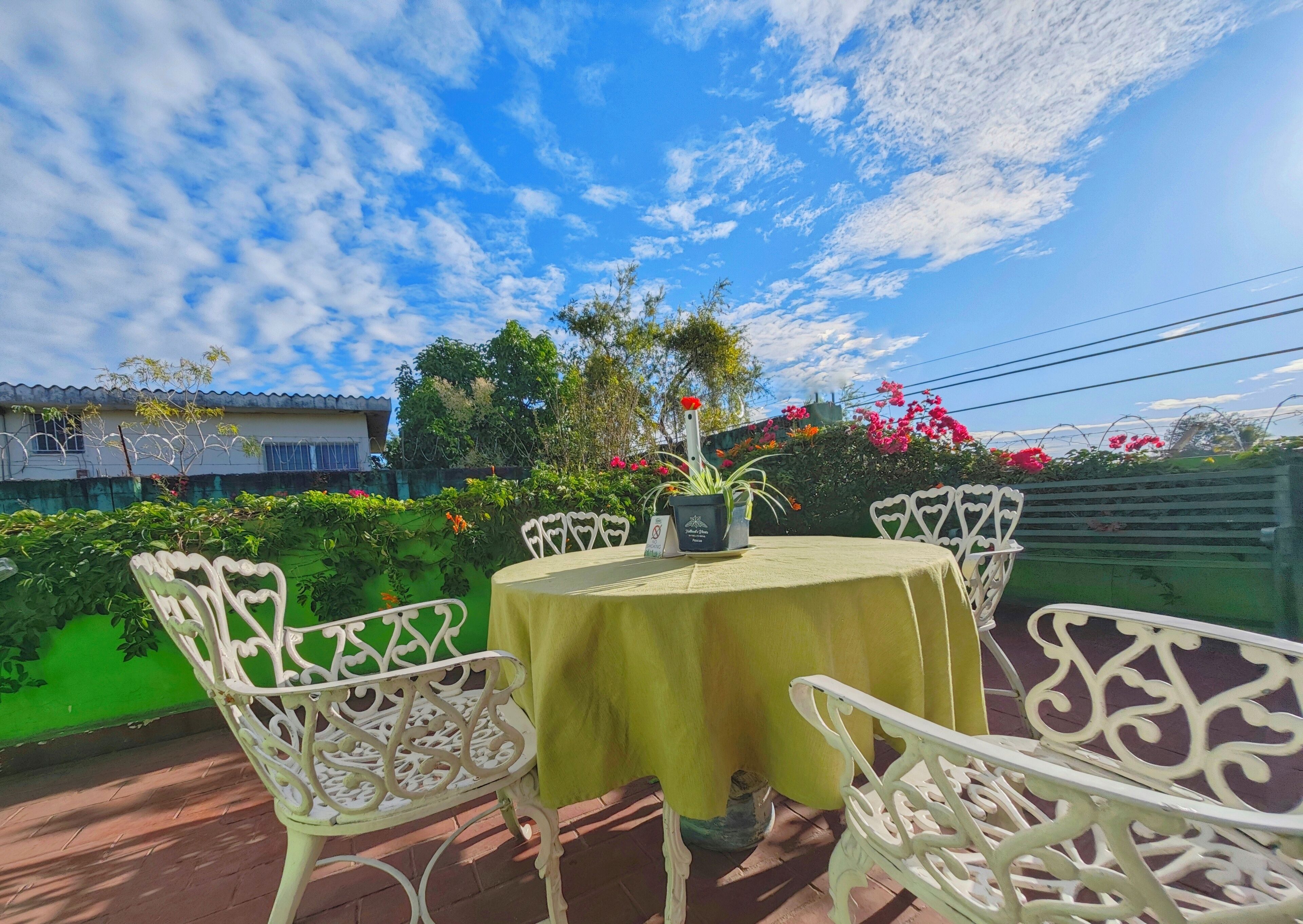 An outdoor patio with a round table and white chairs, surrounded by greenery and flowering plants.