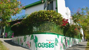 Een hotel genaamd 'oasis' met een gevel in groen en wit, een balkon met planten en een helderblauwe lucht.
