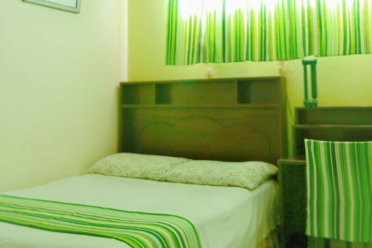 Una habitación con cama, escritorio y silla. Hay un ventilador de techo y una ventana con cortinas.