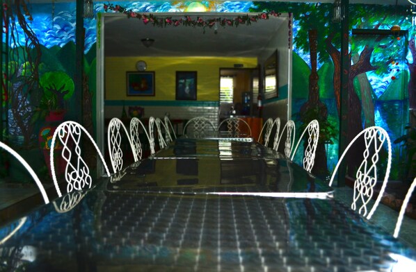 A dining area with a long table and white chairs, surrounded by walls with murals and potted plants. - Hotel Oasis (San Salvador)