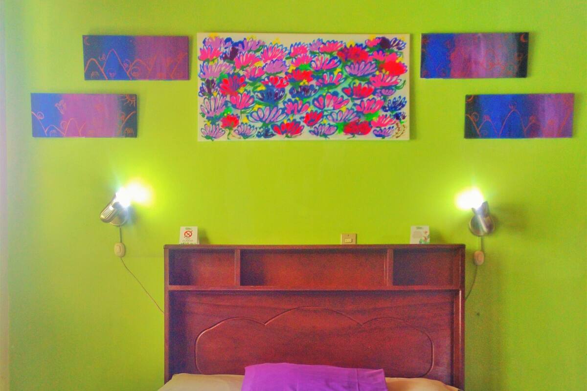 Una cama con cabecera de madera, dos lámparas fijadas a la pared y un cuadro floral colorido encima.