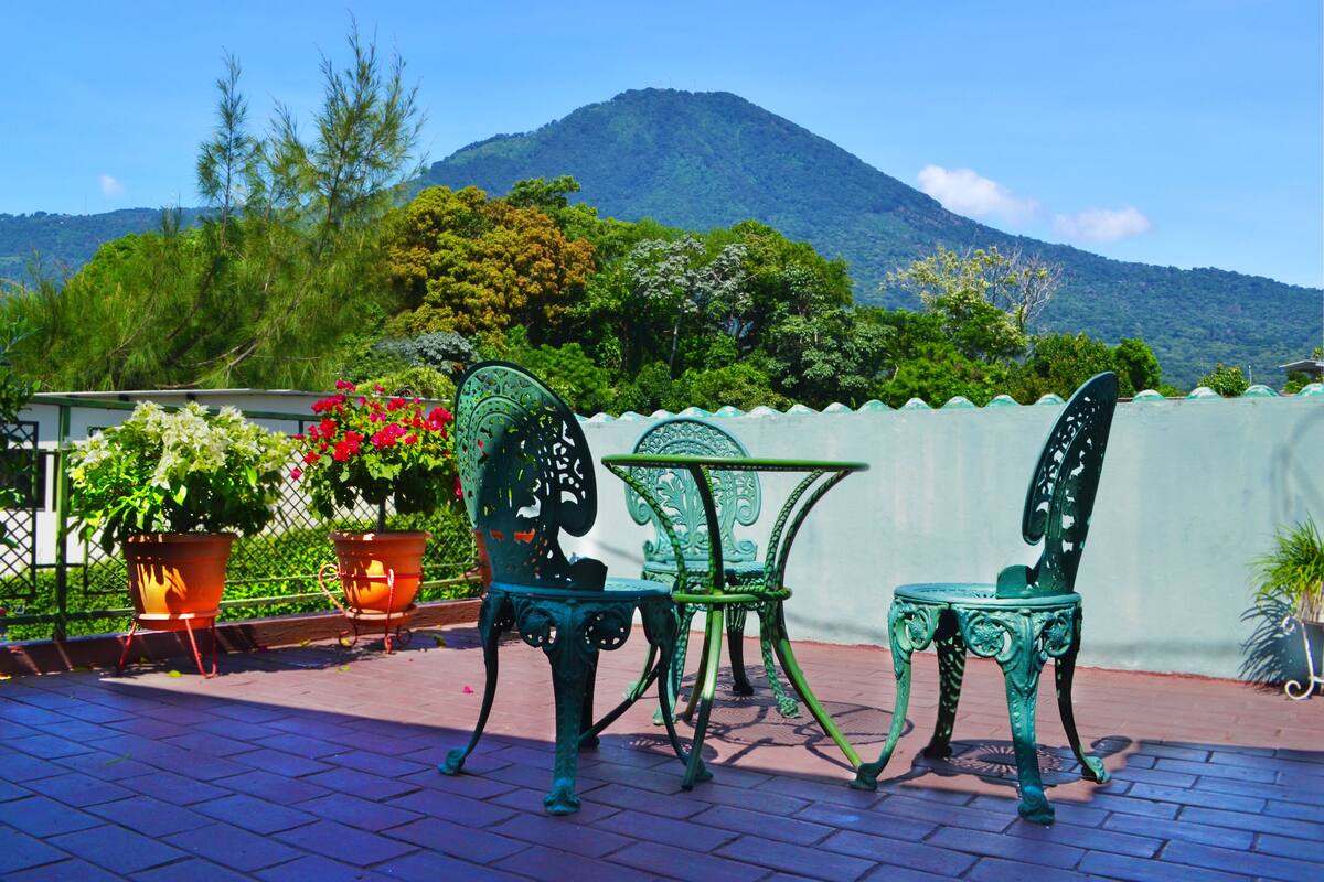 Un patio con dos sillas metálicas verdes y una mesa, rodeado de plantas en macetas y vistas a una montaña.