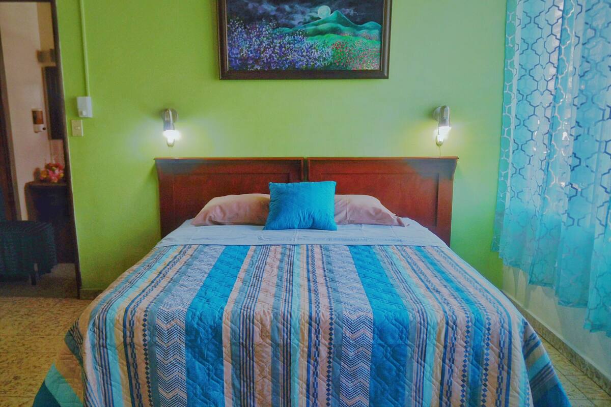 Una habitación con una cama, paredes verdes, un cuadro y una cortina estampada.