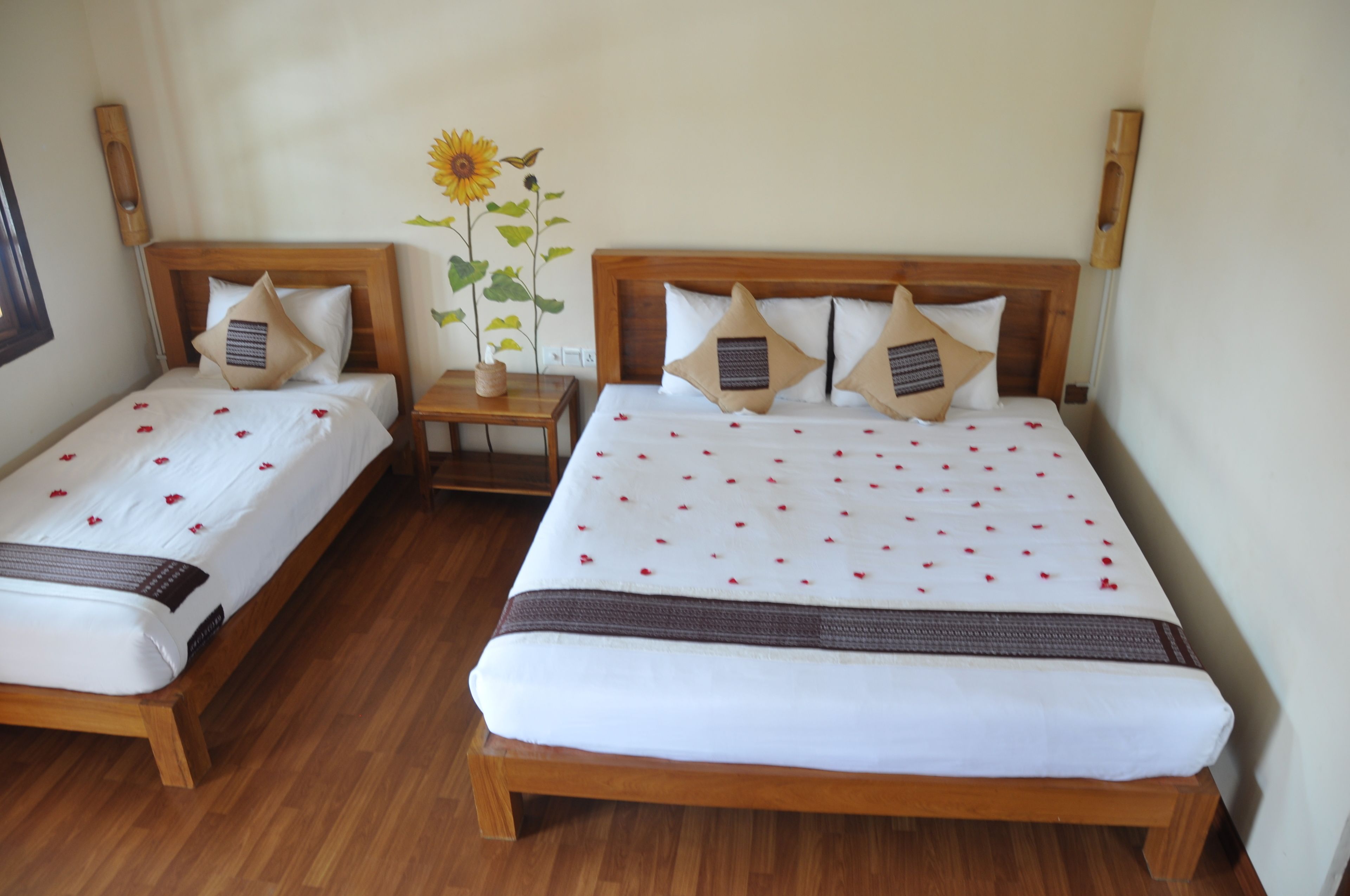 Een kamer met twee eenpersoonsbedden, elk met een houten hoofdbord, wit beddengoed en decoratieve kussens. Daartussen staat een klein houten nachtkastje met een bloemenvaas.