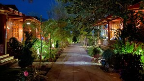 A pathway flanked by buildings with warm lighting, surrounded by lush greenery and potted plants. - Trinity Family Inn (Nyaungshwe)