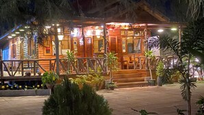 A wooden cabin-style building with a porch and outdoor seating area, surrounded by potted plants and trees. - Trinity Family Inn (Nyaungshwe)