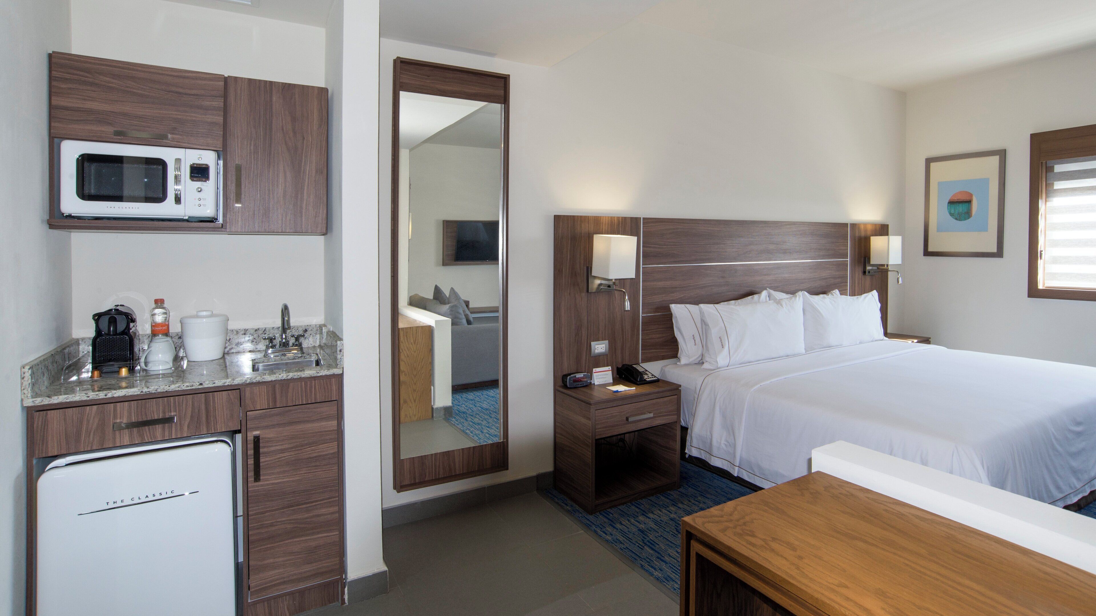 Habitación de hotel con cama, mesita de noche, microondas, refrigerador pequeño, lavamanos y espejo.