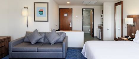 Een hotelkamer met een grijze bank, een wit bed, een kleine kitchenette en een badkamer door een open deur zichtbaar.