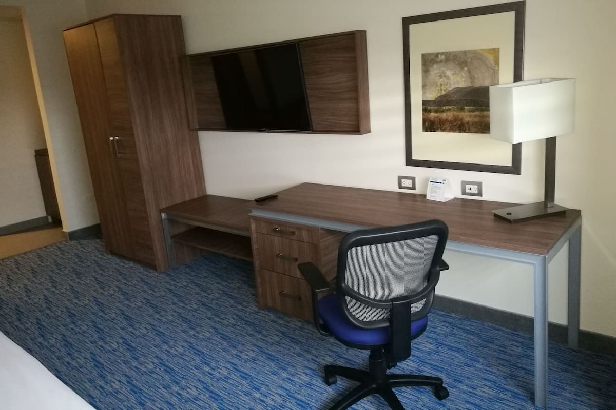 A hotel room with a wooden desk, a blue carpet, a black office chair, a TV mounted on the wall, and a framed picture above the desk.