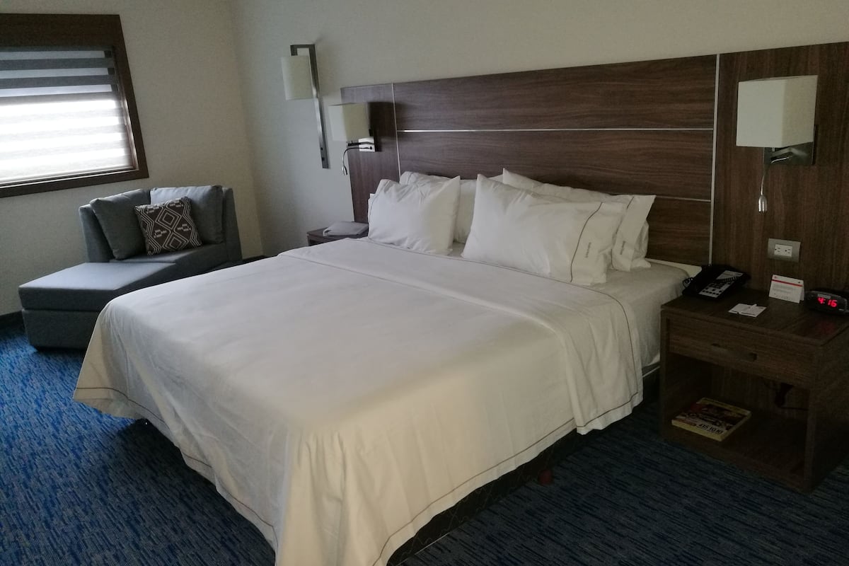 A hotel room with a large bed, a bedside table with a phone and a clock, a small seating area with a couch and pillows, and a window with blinds.