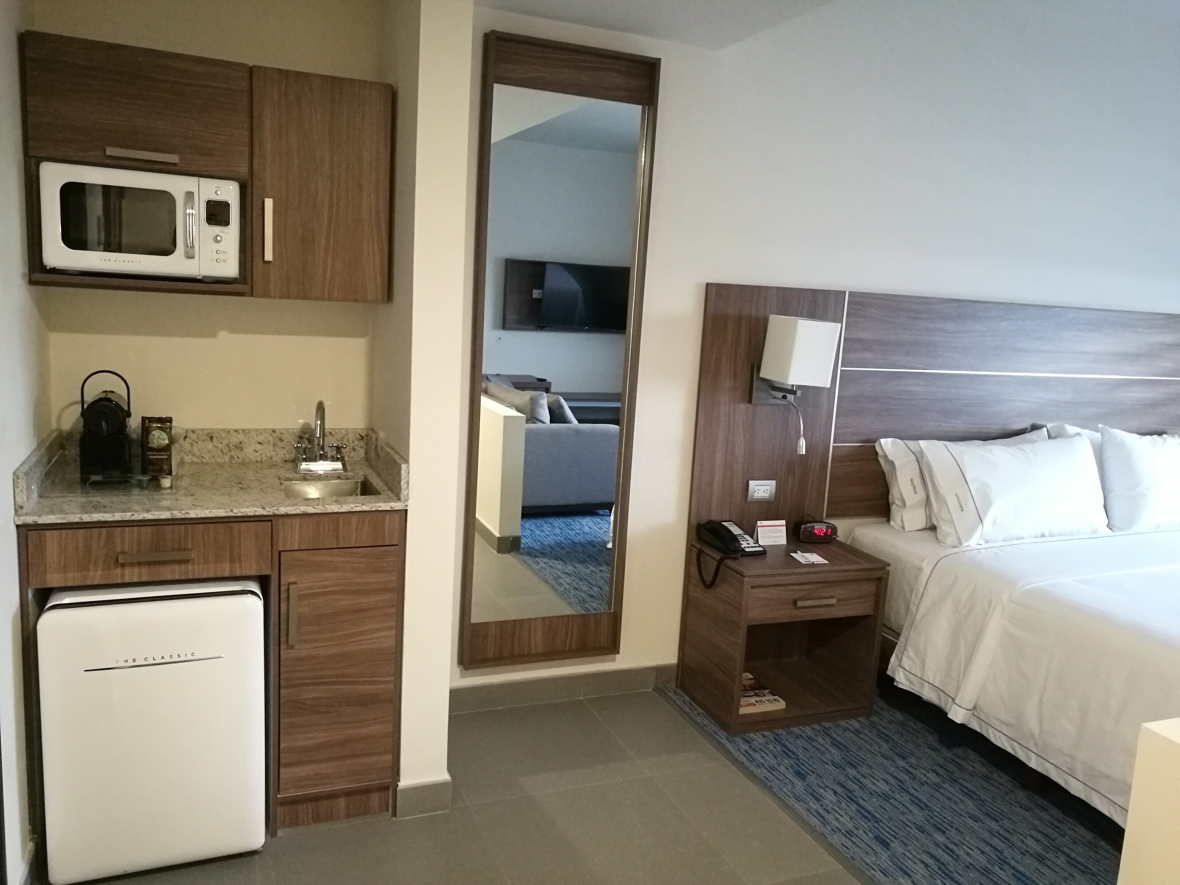 A hotel room with a bed, a bedside table with a lamp, a microwave, a coffee maker, and a mini-fridge.