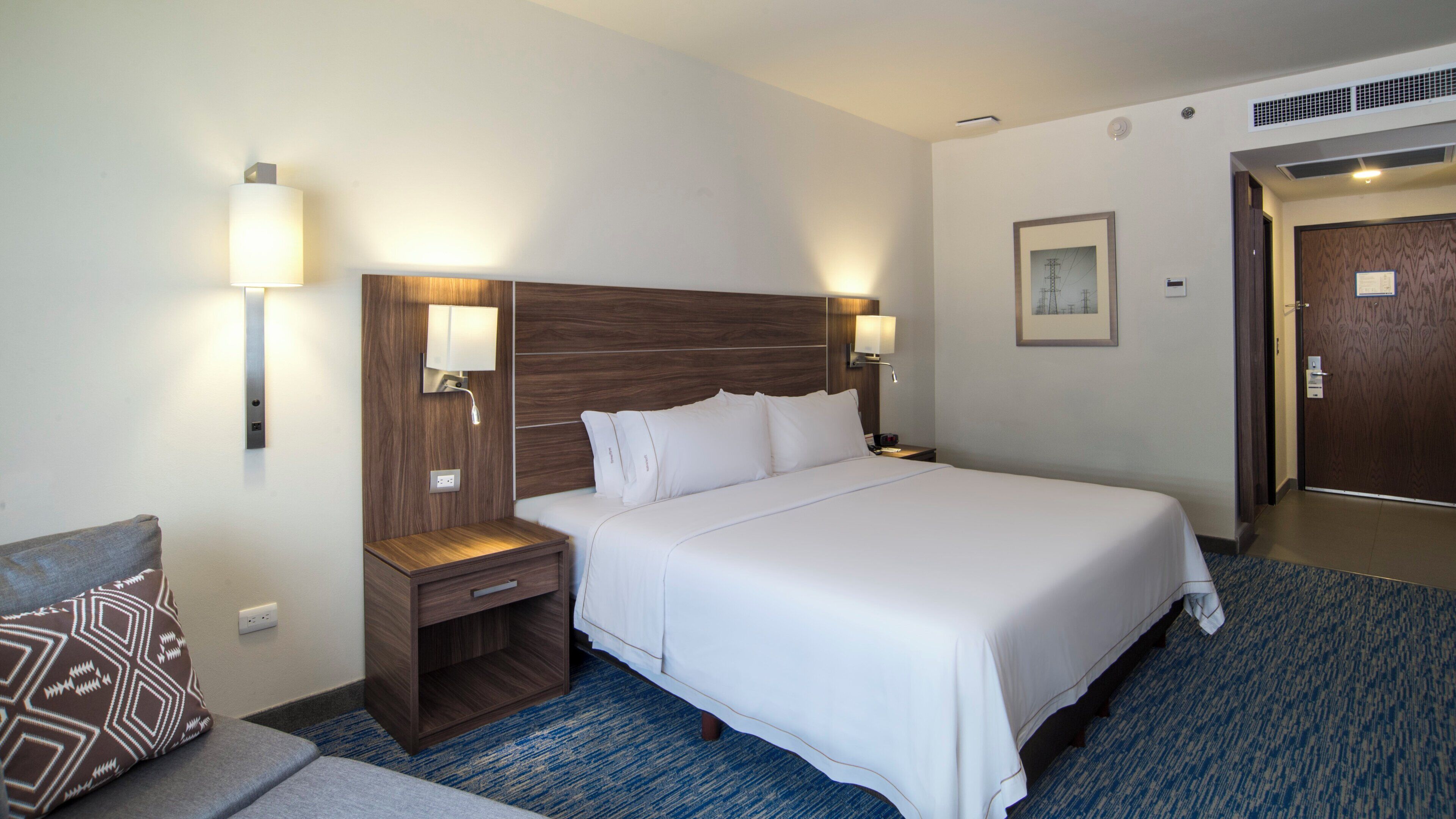 A hotel room with a large bed, bedside tables, wall-mounted lamps, a carpeted floor, and a door.