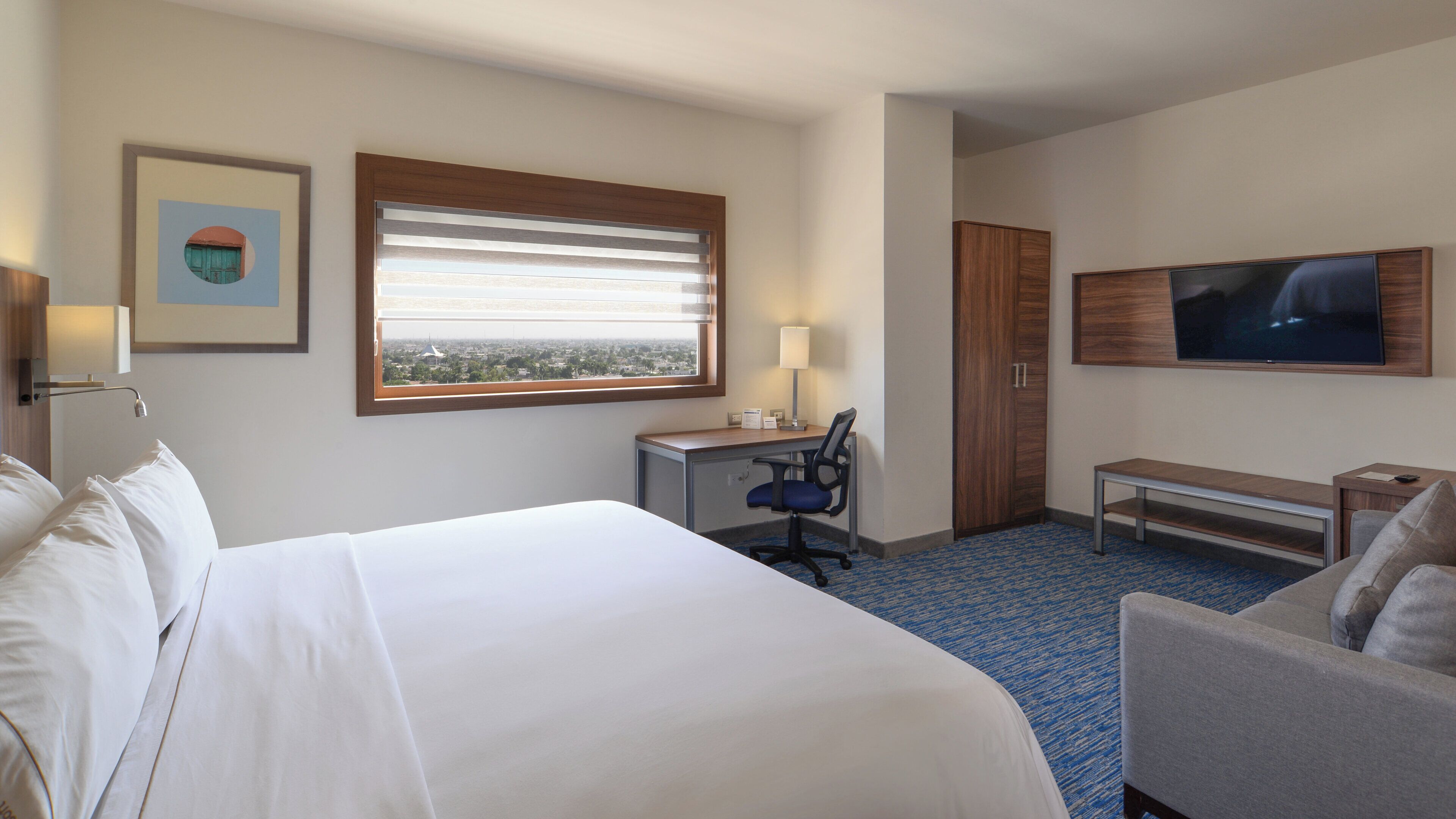 A hotel room with a large bed, a desk with a chair, a television, and a window with blinds.
