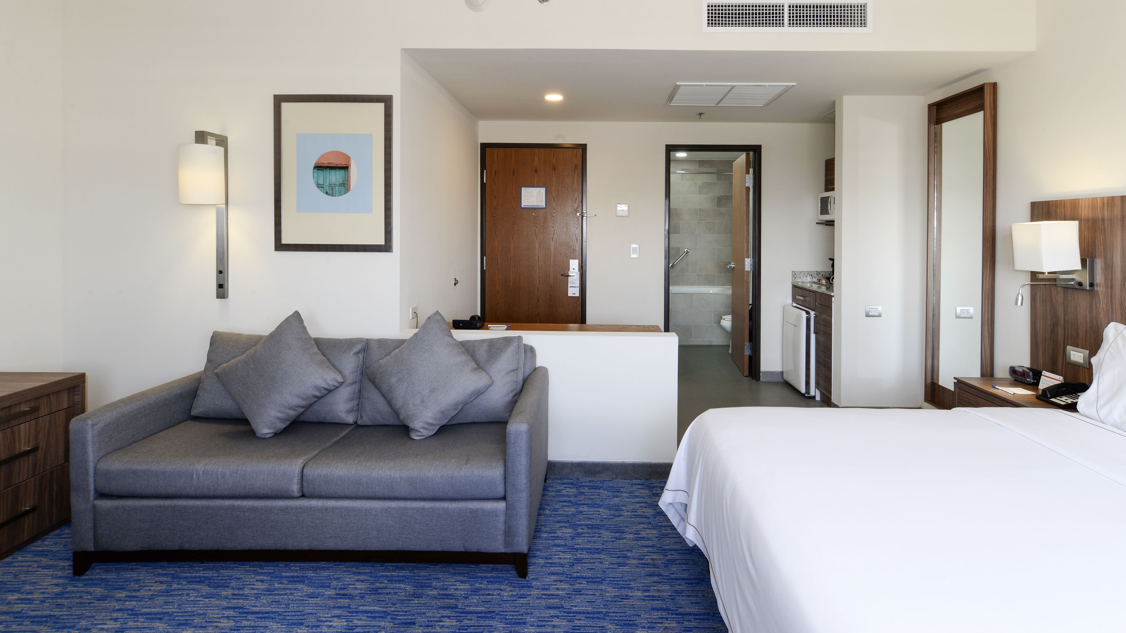A hotel room with a gray sofa, a white bed, a small kitchenette, and a bathroom visible through an open door.