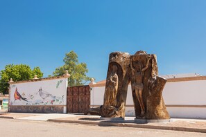 Une sculpture représentant le tronc d’un arbre orné d’une figure sculptée, une fresque murale avec des oiseaux et des fleurs, et un ciel d’un bleu limpide.