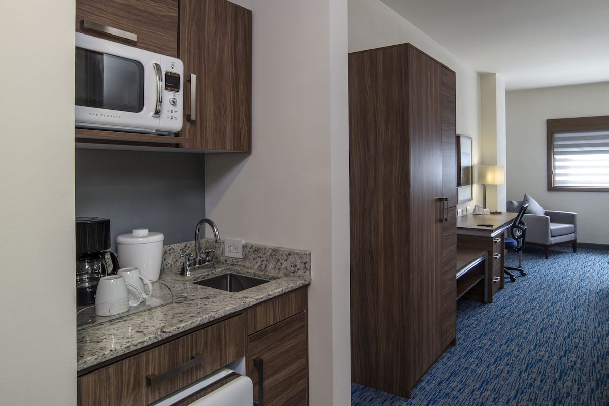 A hotel room with a kitchenette, a bed, a desk, and a window with blinds.
