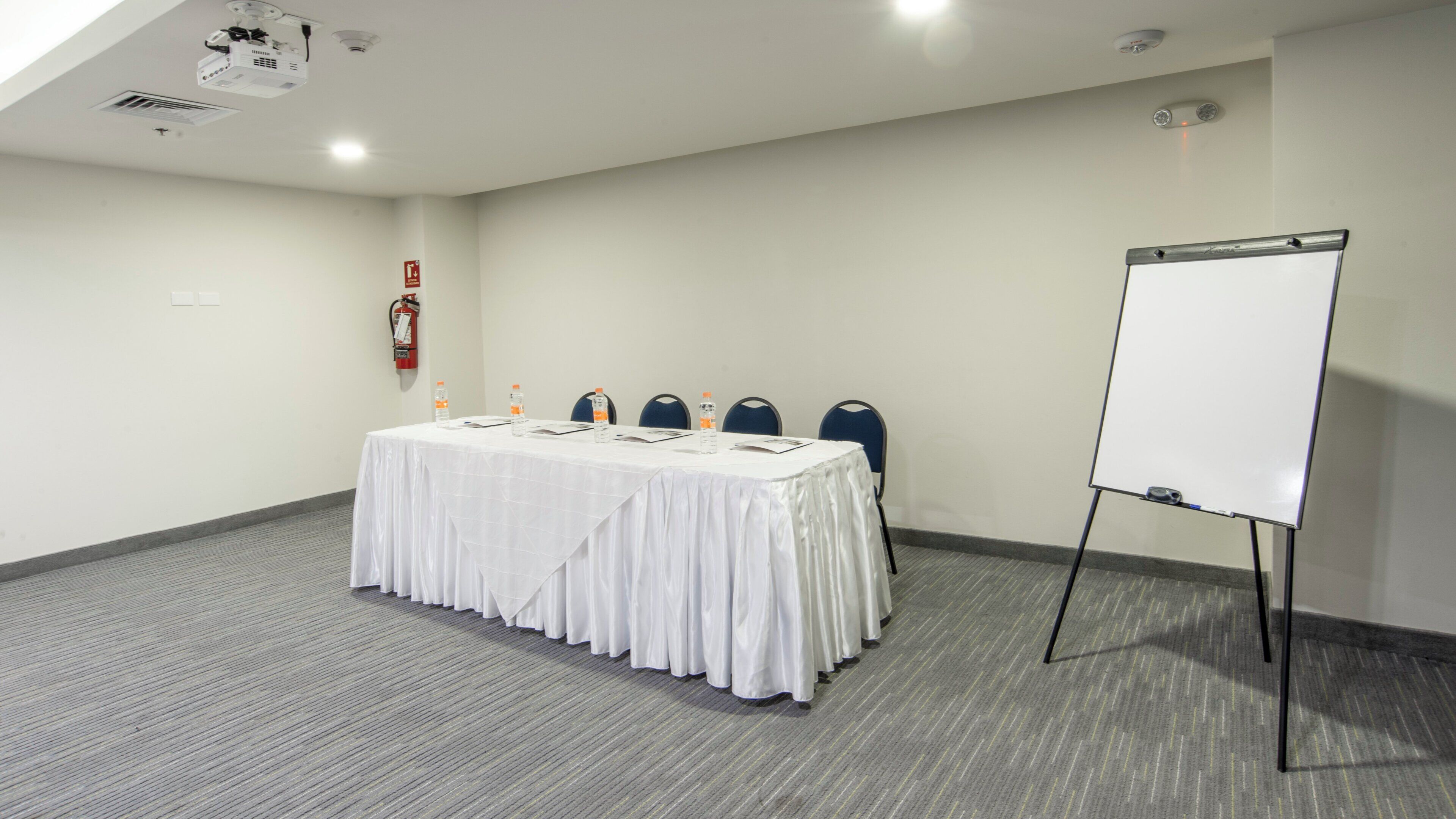 A conference room with a white table, chairs, a whiteboard, and a projector on the ceiling.