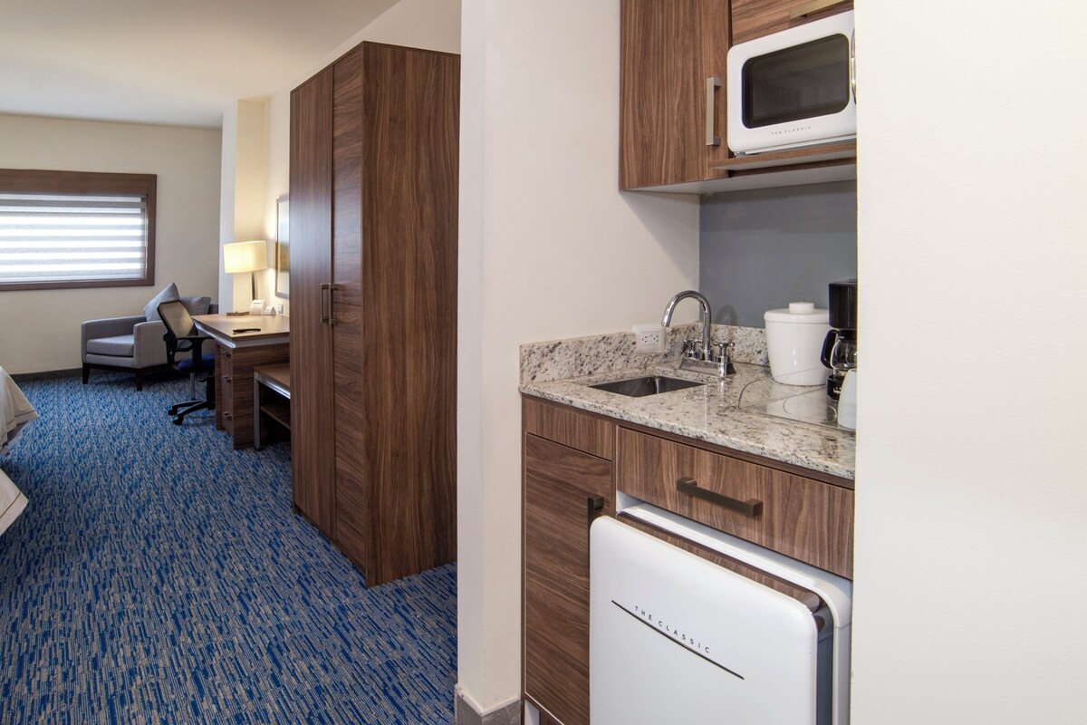 Une chambre d’hôtel équipée d’un lit, d’un bureau, d’une chaise, d’une kitchenette et d’une fenêtre avec des stores.