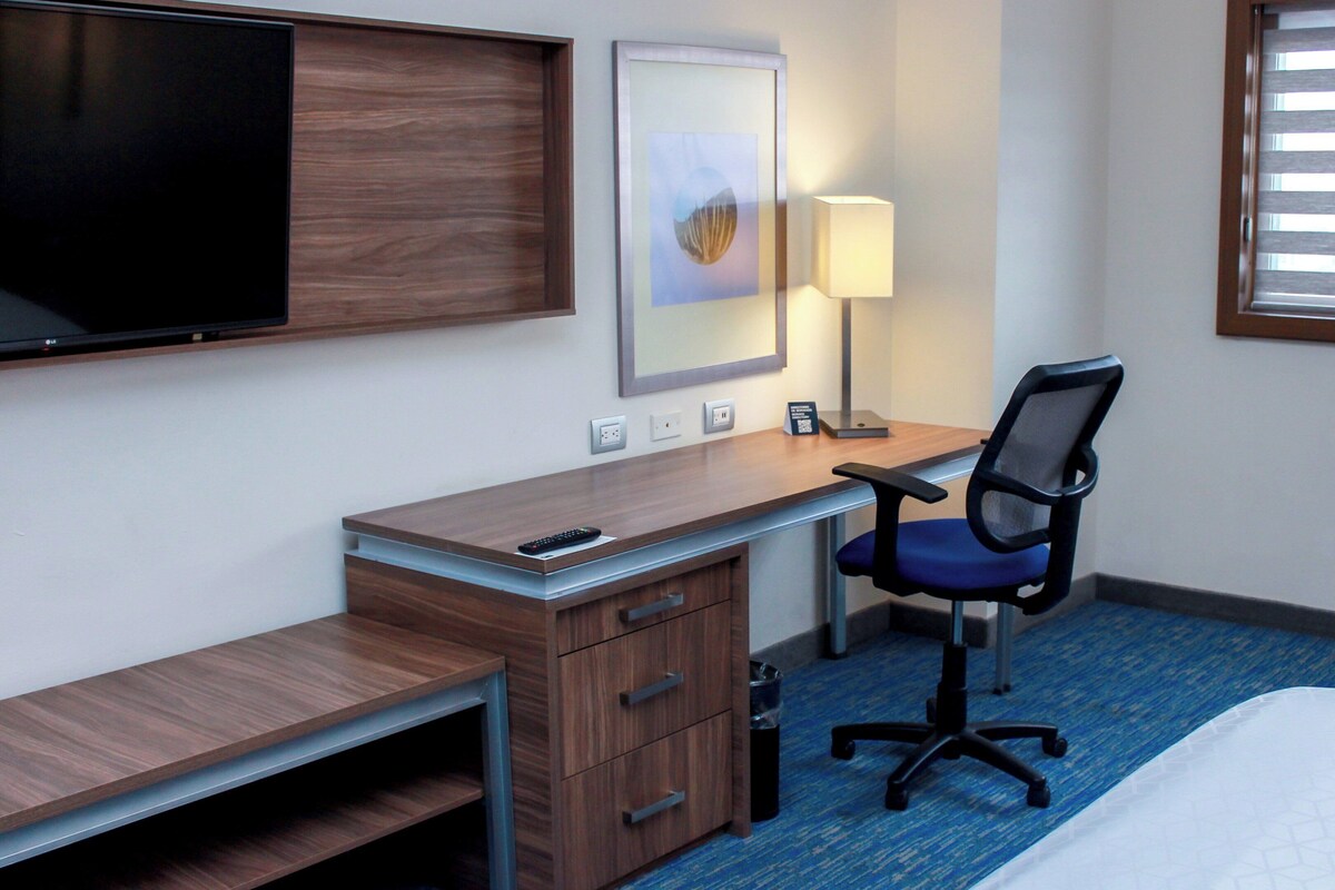 Une chambre d’hôtel avec un bureau en bois, une chaise, un lit, une télévision, une lampe et une fenêtre avec des stores.