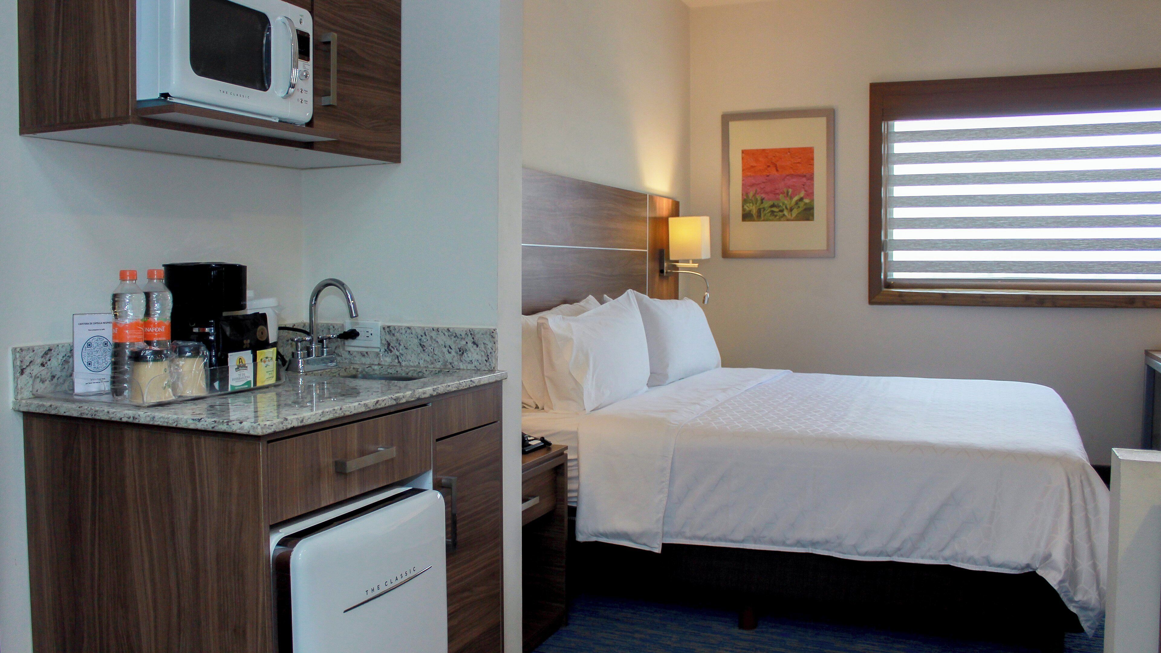 A hotel room with a kitchenette, a bed, a nightstand, a lamp, and a window with blinds.
