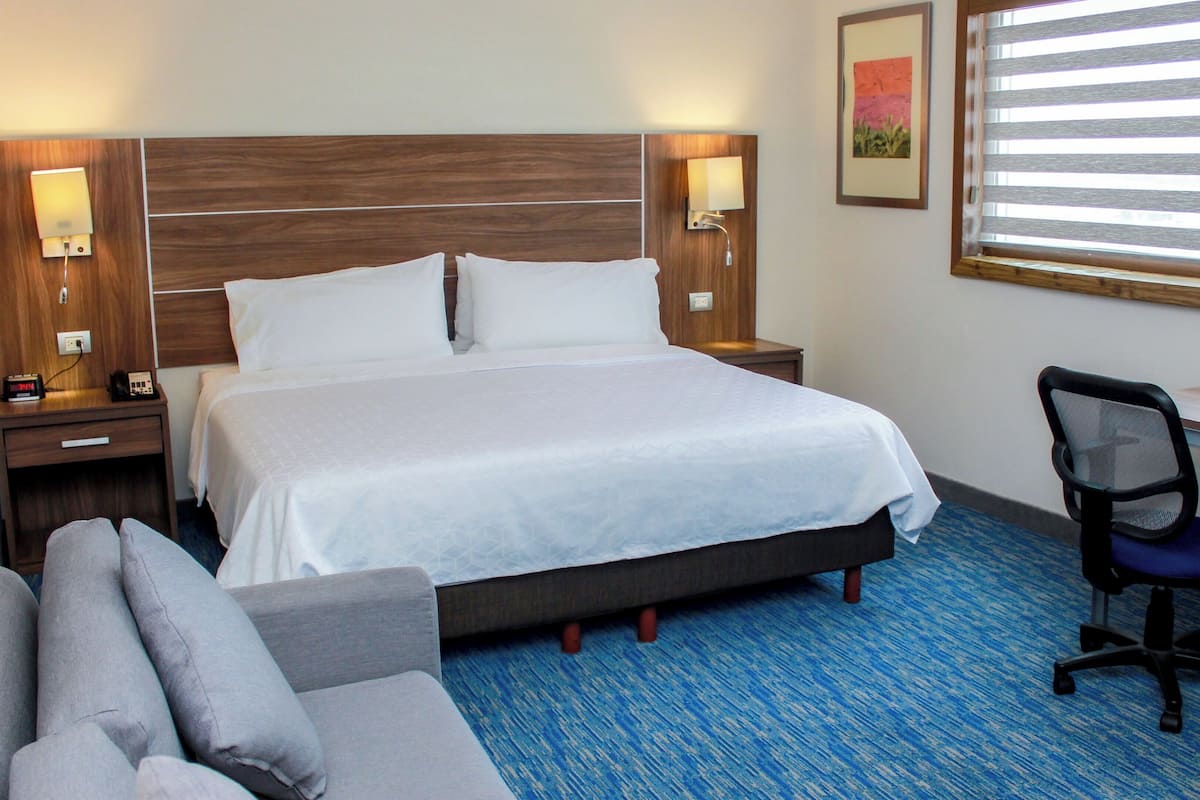A hotel room with a large bed, bedside lamps, a desk, a chair, and a window with blinds.