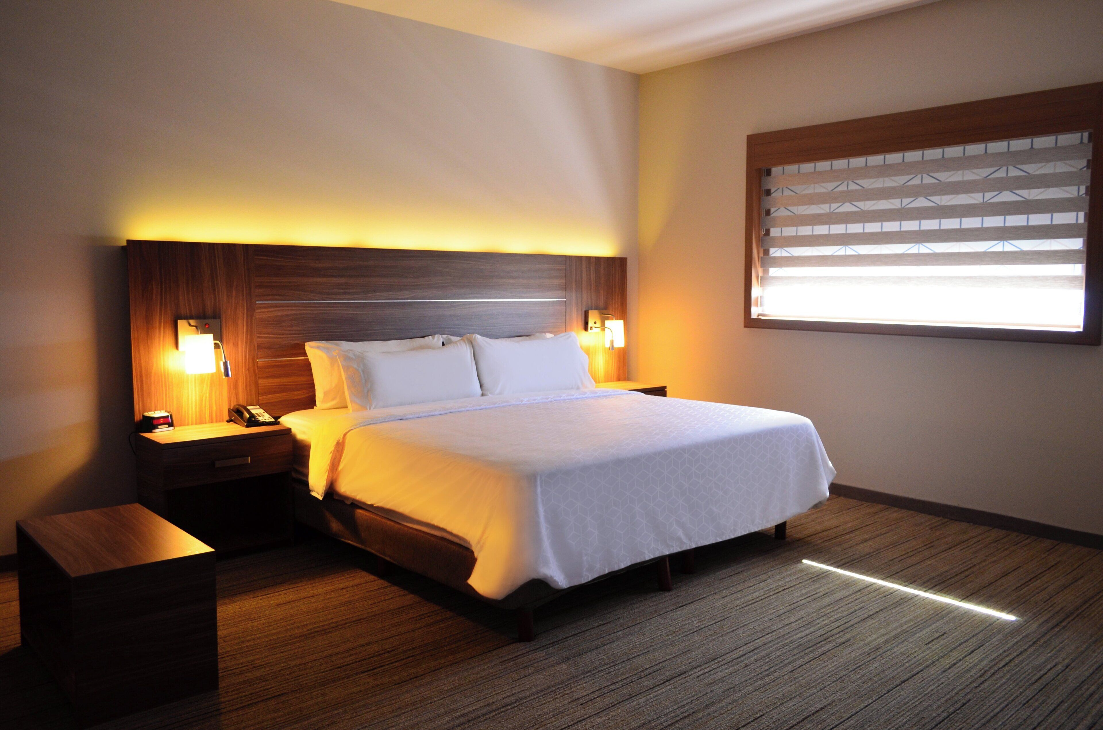 A hotel room with a bed, bedside tables, a nightstand, a lamp, and a window with blinds.