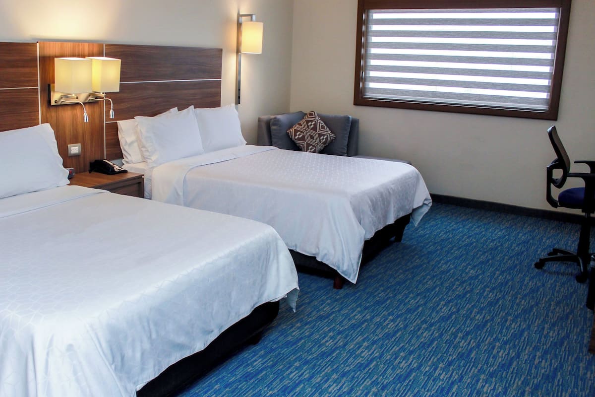 A hotel room with two beds, a nightstand with a lamp, a chair, and a window with blinds.