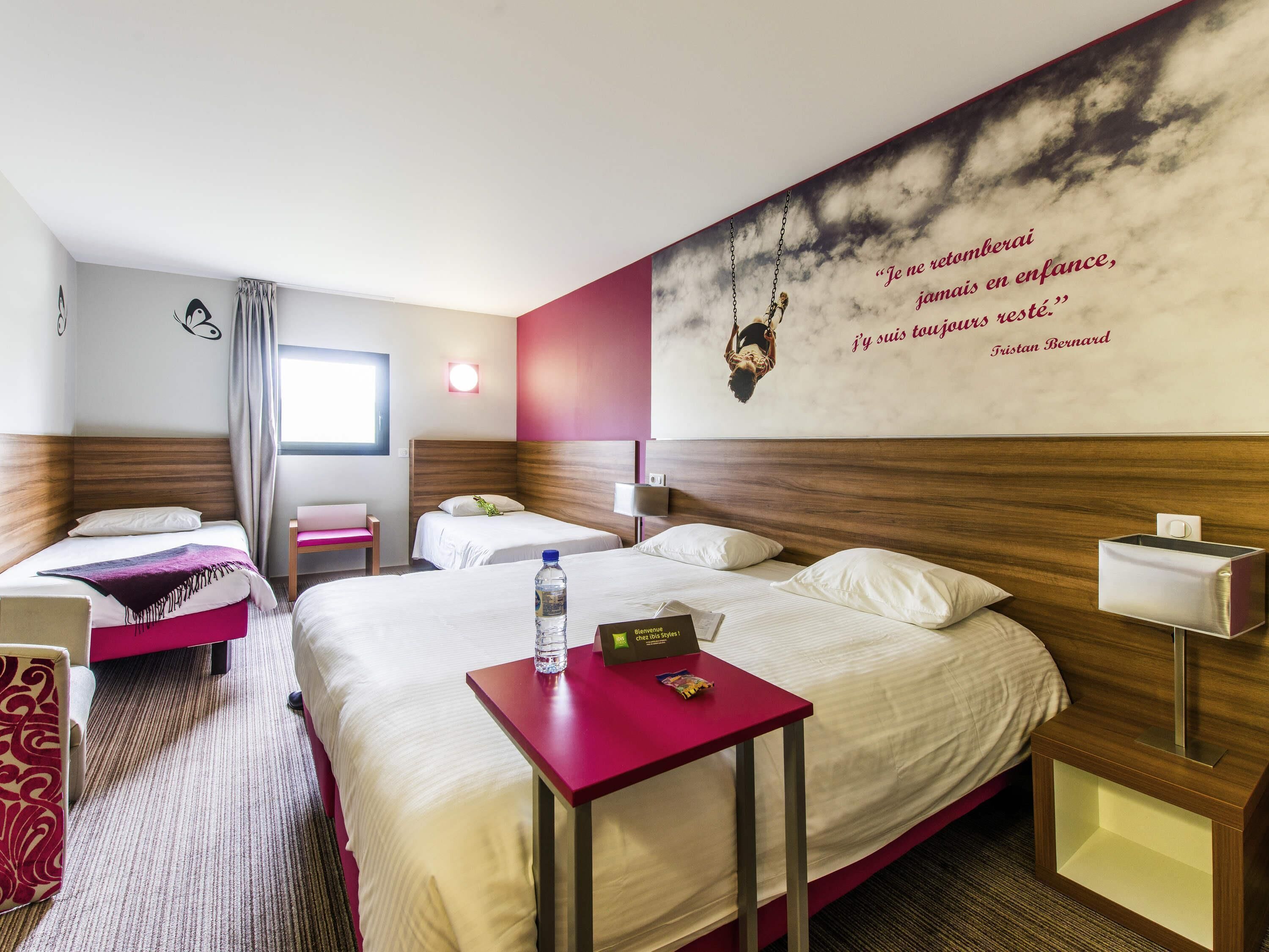 A hotel room with two beds, a red table, a wall clock, and a quote by Tristan Bernard.